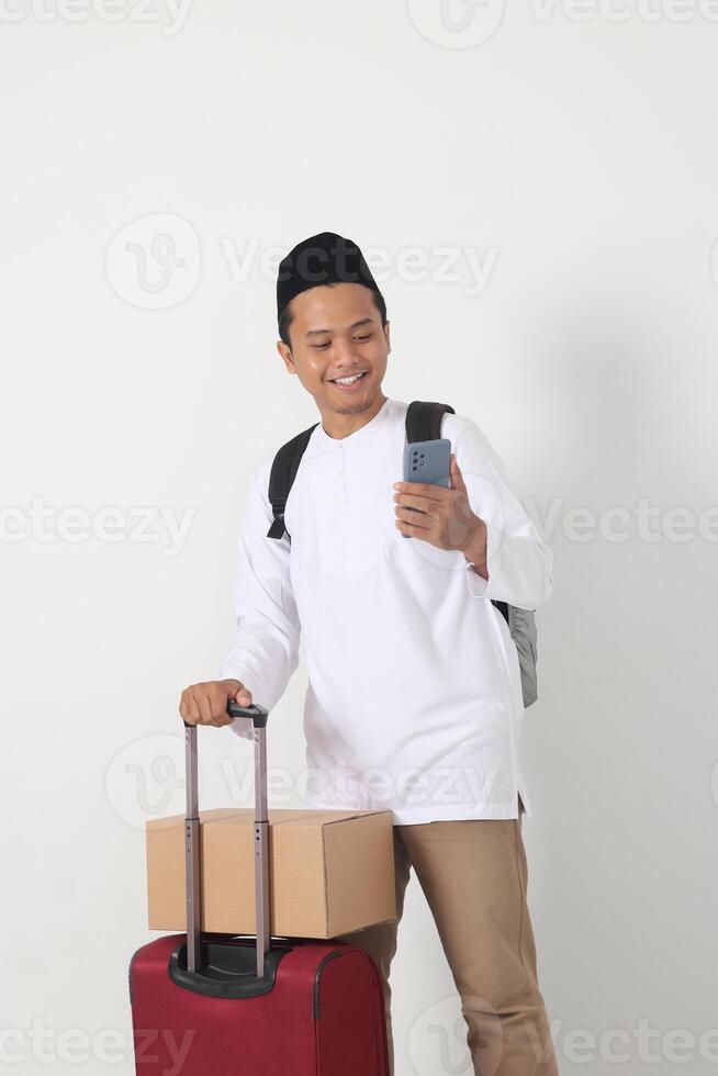 Portrait of happy Asian muslim man carrying cardboard box and suitcase while holding mobile phone. Going home for Eid Mubarak. Isolated image on white background photo
