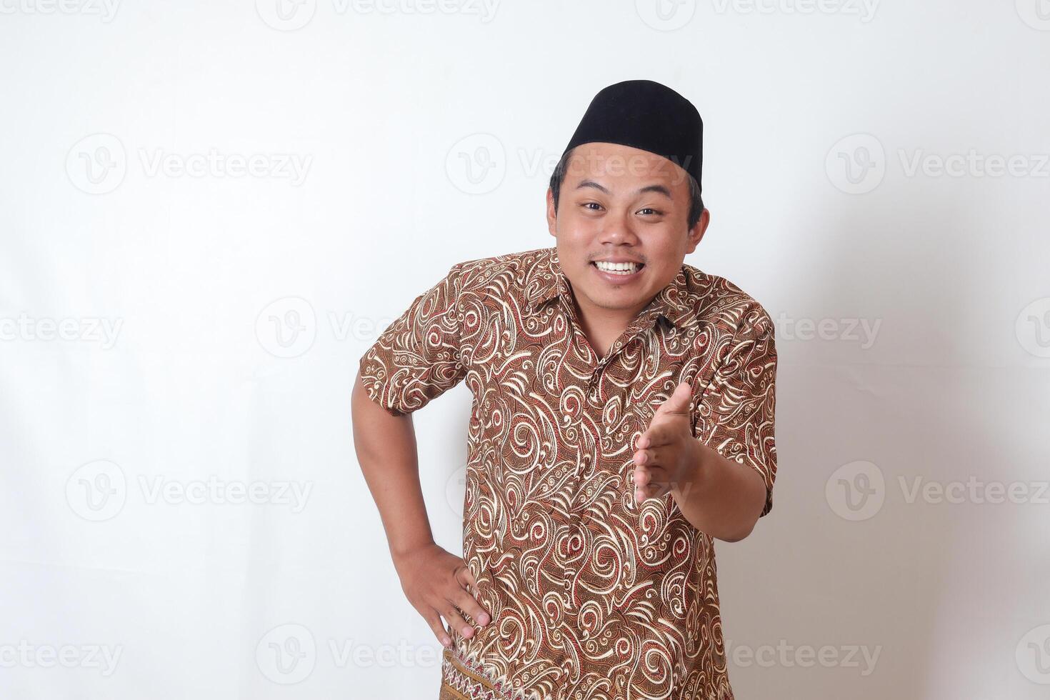 retrato de emocionado asiático hombre vistiendo batik camisa y Songkok alcanzando fuera su mano para un mano agitar, acogedor alguien. aislado imagen en gris antecedentes foto
