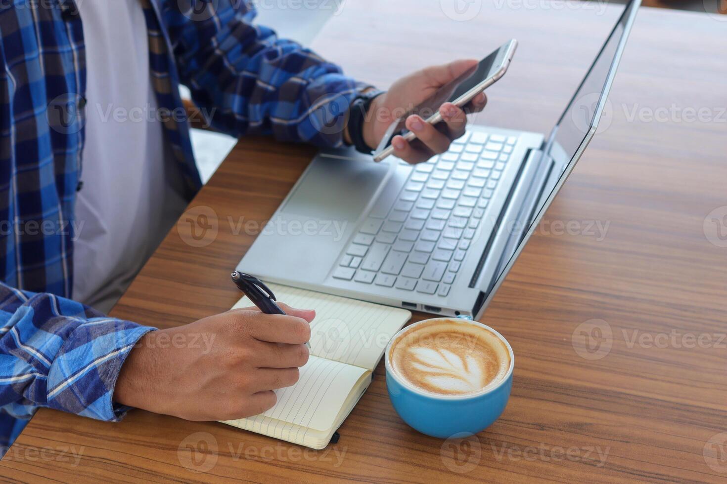 Close up of male hand writing on note book with pen, showing blank screen of laptop and smartphone. Working in cafe concept with a cup of coffee photo