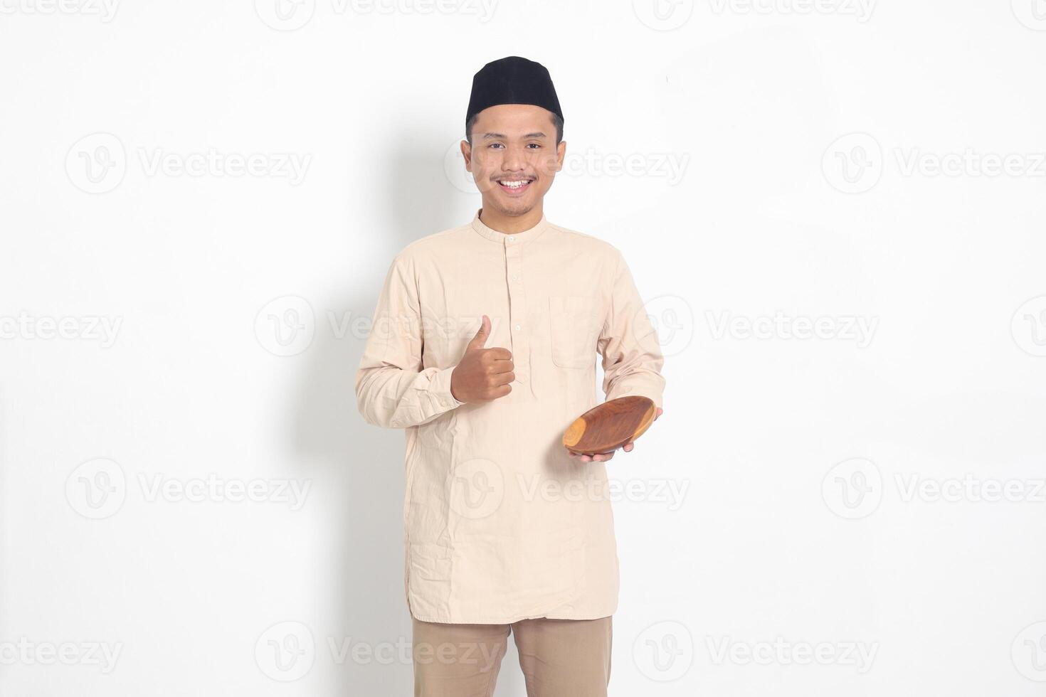 Portrait of excited Asian muslim man in koko shirt with skullcap showing and holding an empty plate. Bowl template for food brand. Isolated on white background photo