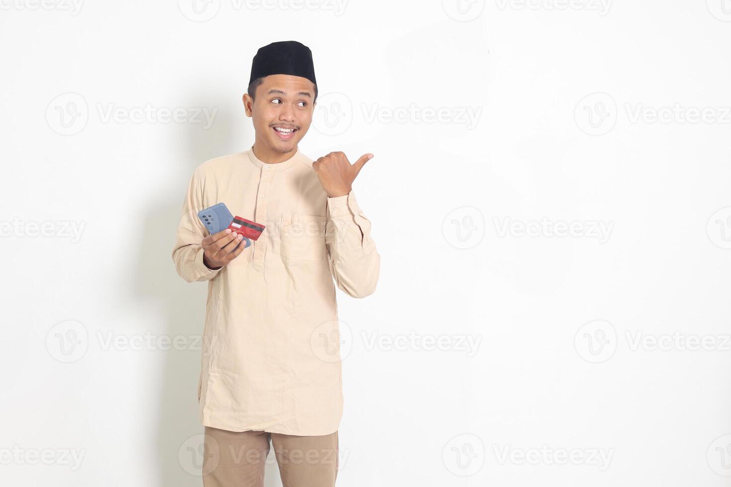 retrato de atractivo asiático musulmán hombre en koko camisa con casquete participación un móvil teléfono y crédito tarjeta mientras señalando con dedo a el lado. aislado imagen en blanco antecedentes foto