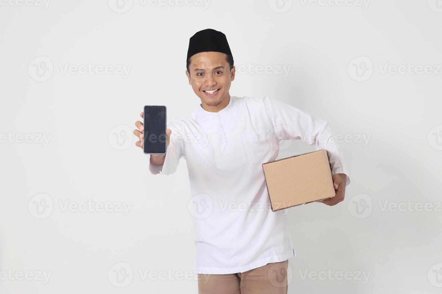 Portrait of happy Asian muslim man in koko shirt with peci carrying cardboard box while showing blank screen mobile phone for mock up. Going home for Eid Mubarak. Isolated image on white background photo