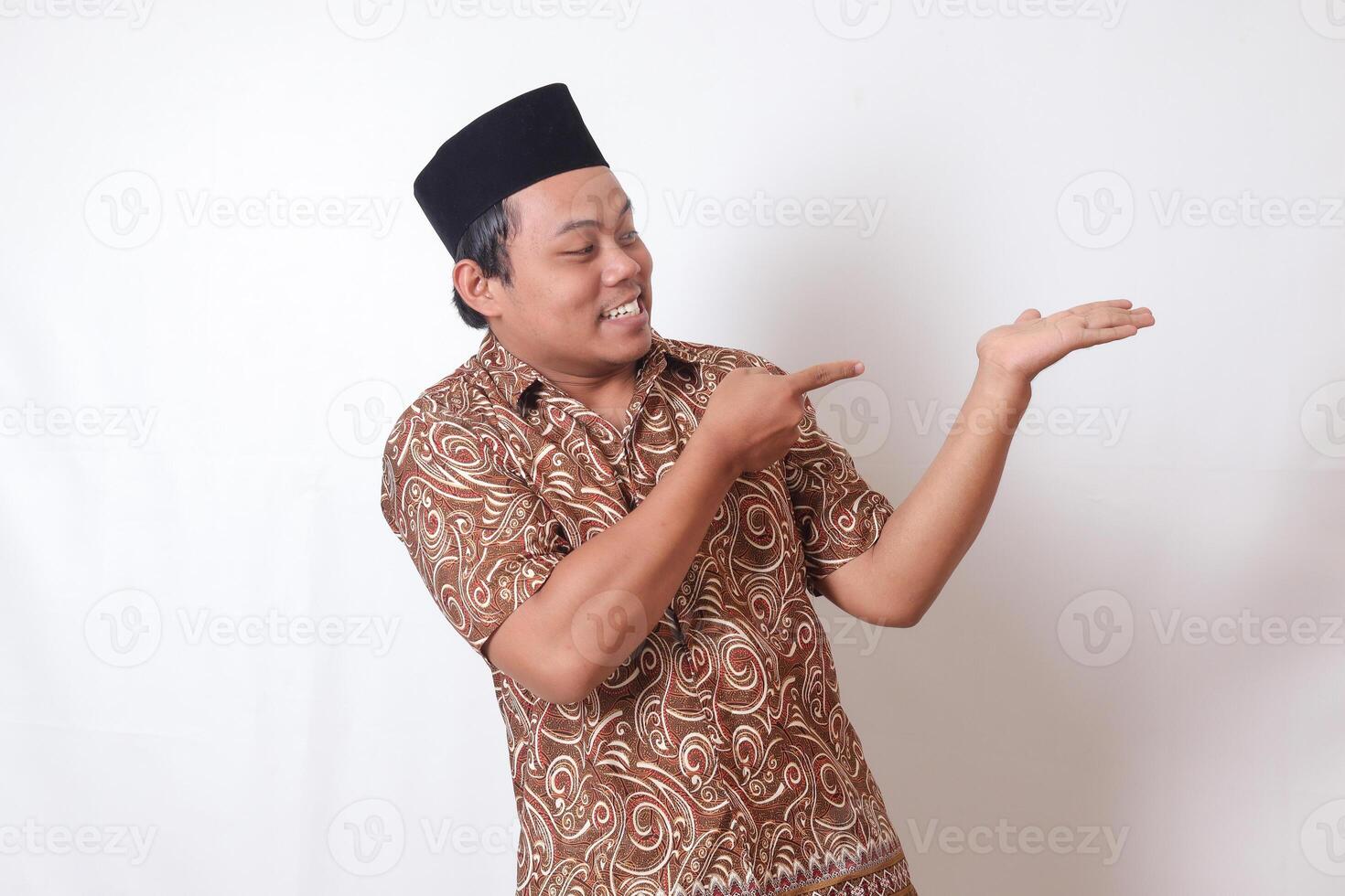 Portrait of excited Asian man wearing batik shirt and songkok smiling and looking at the camera pointing with two hands and fingers to the side. Isolated image on gray background photo