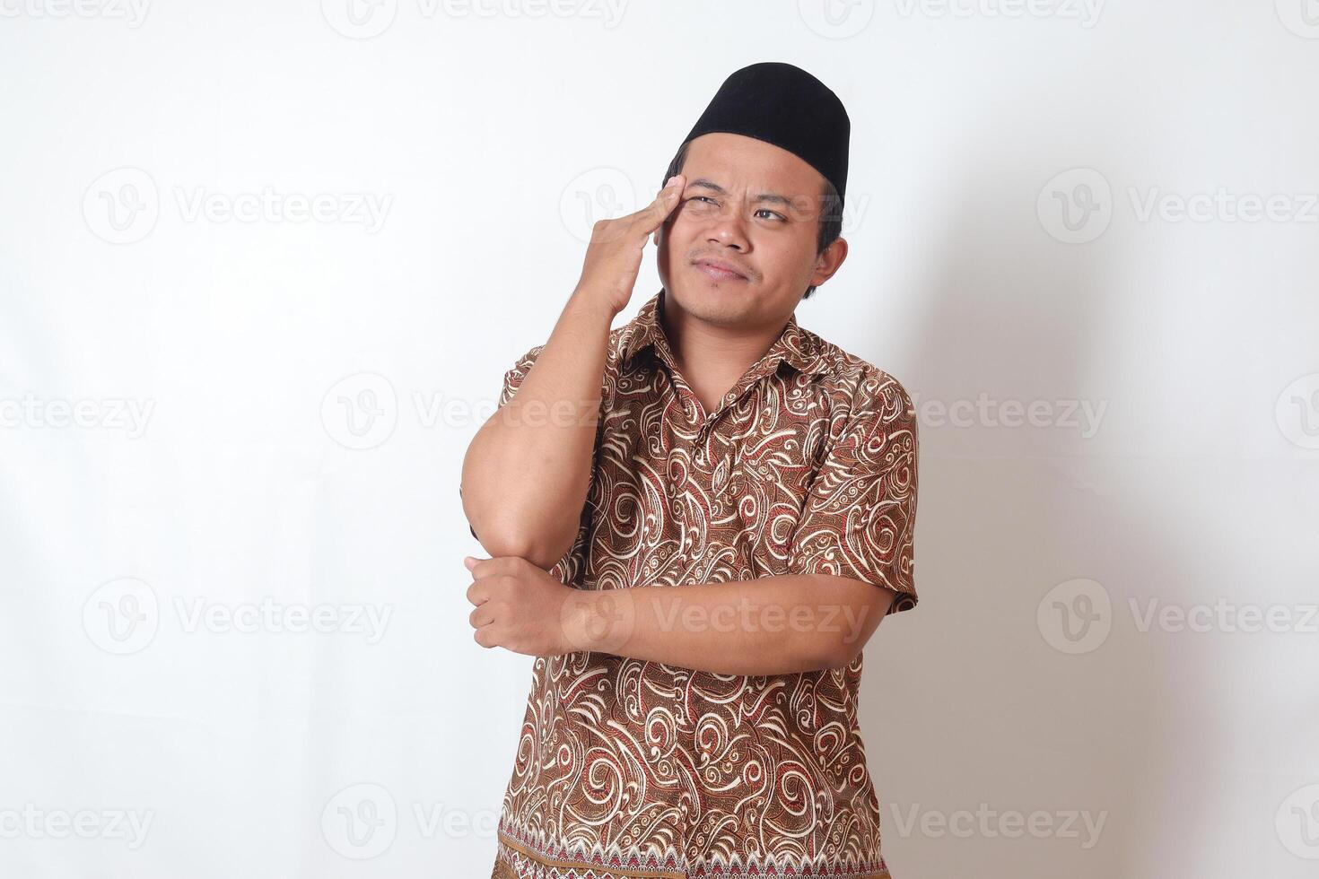 Portrait of suffering Asian man wearing batik shirt and songkok having headache while touching his forehead area. Isolated image on gray background photo