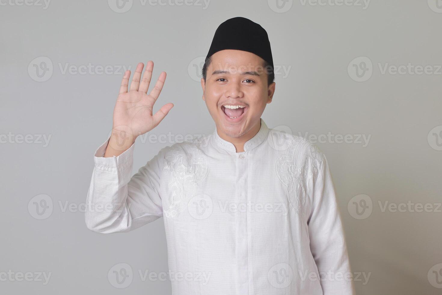 Portrait of Asian muslim man in white koko shirt with skullcap shirt saying hi, waving hand at camera, greeting person with happy smile. Isolated image on white background photo