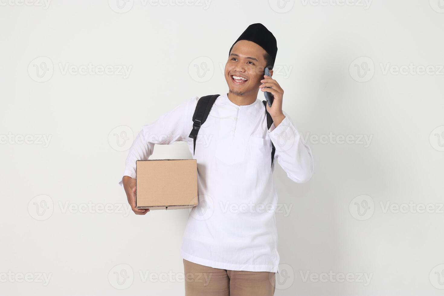 Portrait of happy Asian muslim man in koko shirt with peci carrying cardboard box while talking to his friend and family on cellphone. Going home for Eid Mubarak. Isolated image on white background photo