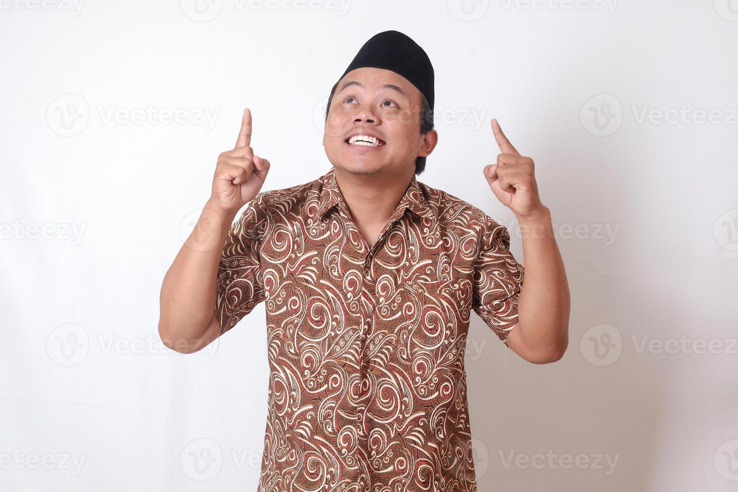 retrato de emocionado asiático hombre vistiendo batik camisa y Songkok sonriente y mirando a el cámara señalando con dos manos y dedos a arriba. aislado imagen en gris antecedentes foto