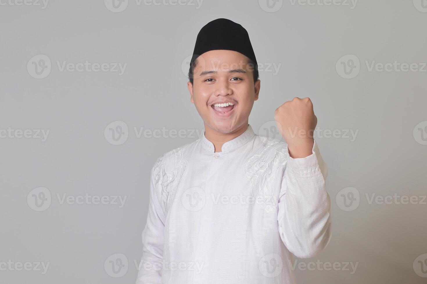 Portrait of Asian muslim man in white koko shirt with skullcap raising his fist, celebrating success. Isolated image on gray background photo