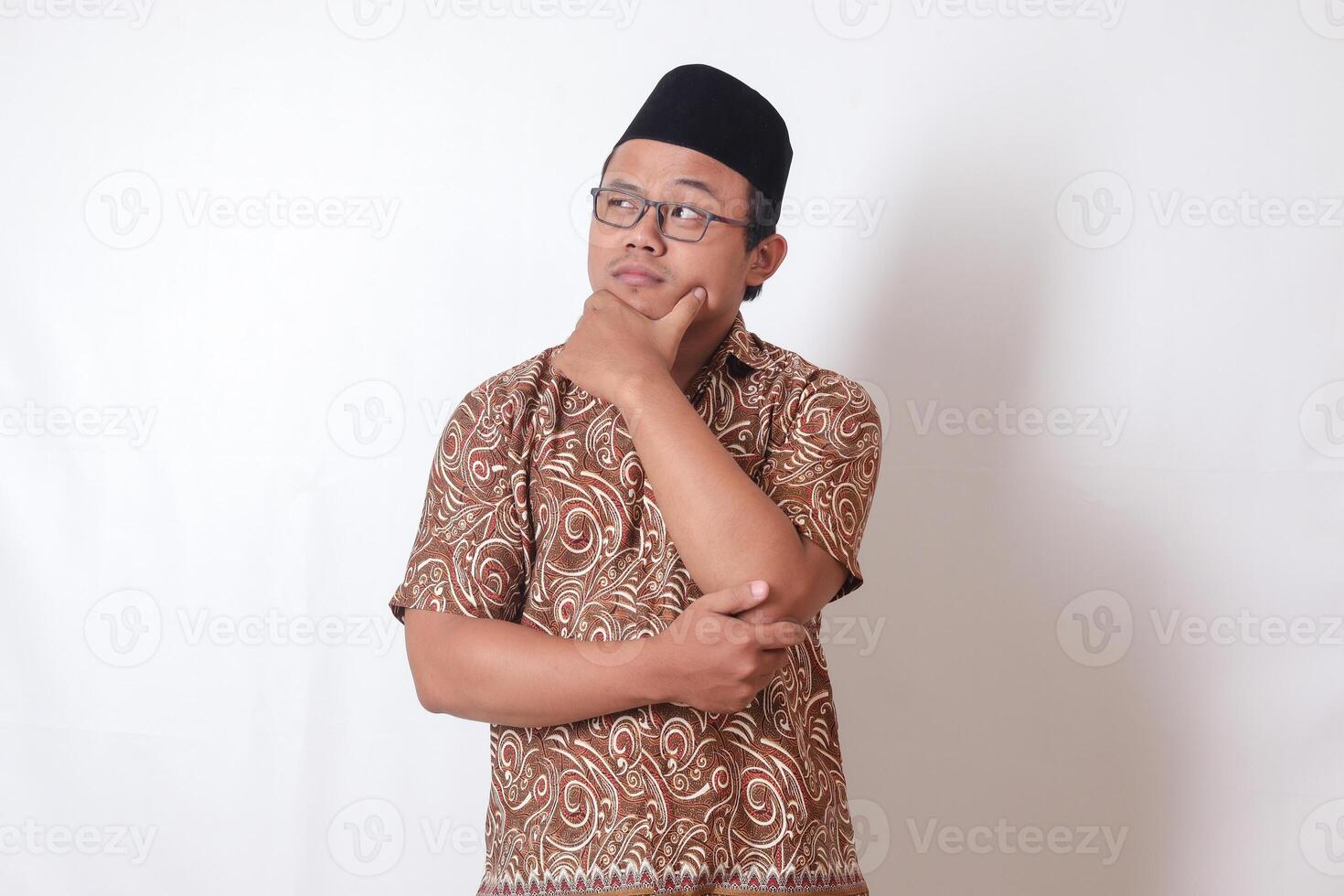 Portrait of confused Asian man wearing batik shirt and songkok standing against gray background, thinking about question with hand on chin photo