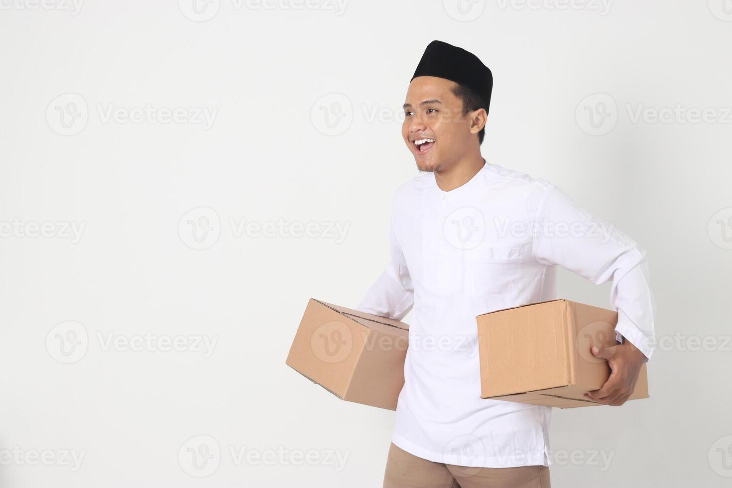 Portrait of happy Asian muslim man in koko shirt with peci looking confident while carrying cardboard box. Homecoming on Eid Mubarak concept. Isolated image on white background photo