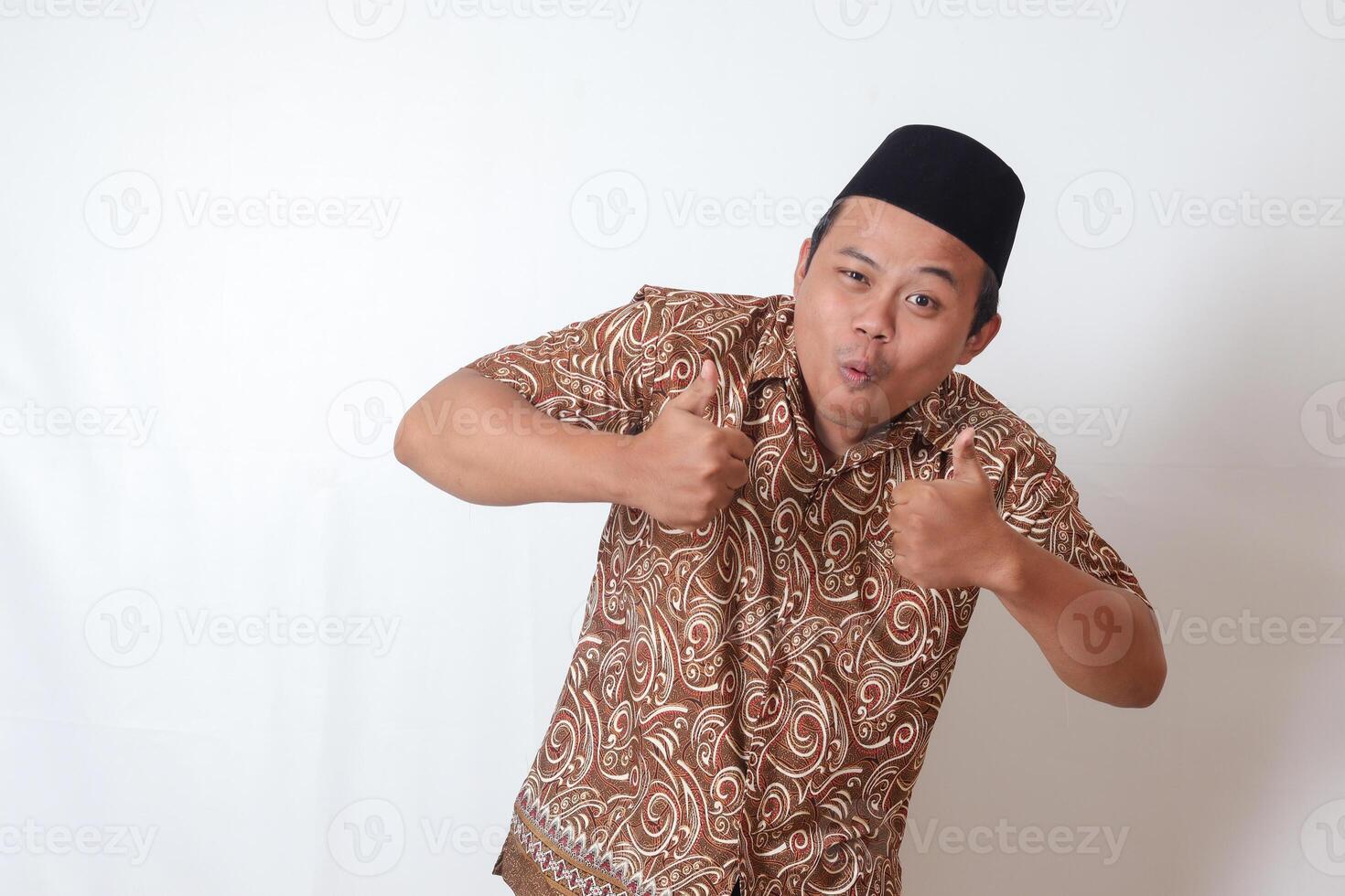 Portrait of excited Asian man wearing batik shirt and songkok smiling and looking at camera, making thumbs up hand gesture. Isolated image on gray background photo