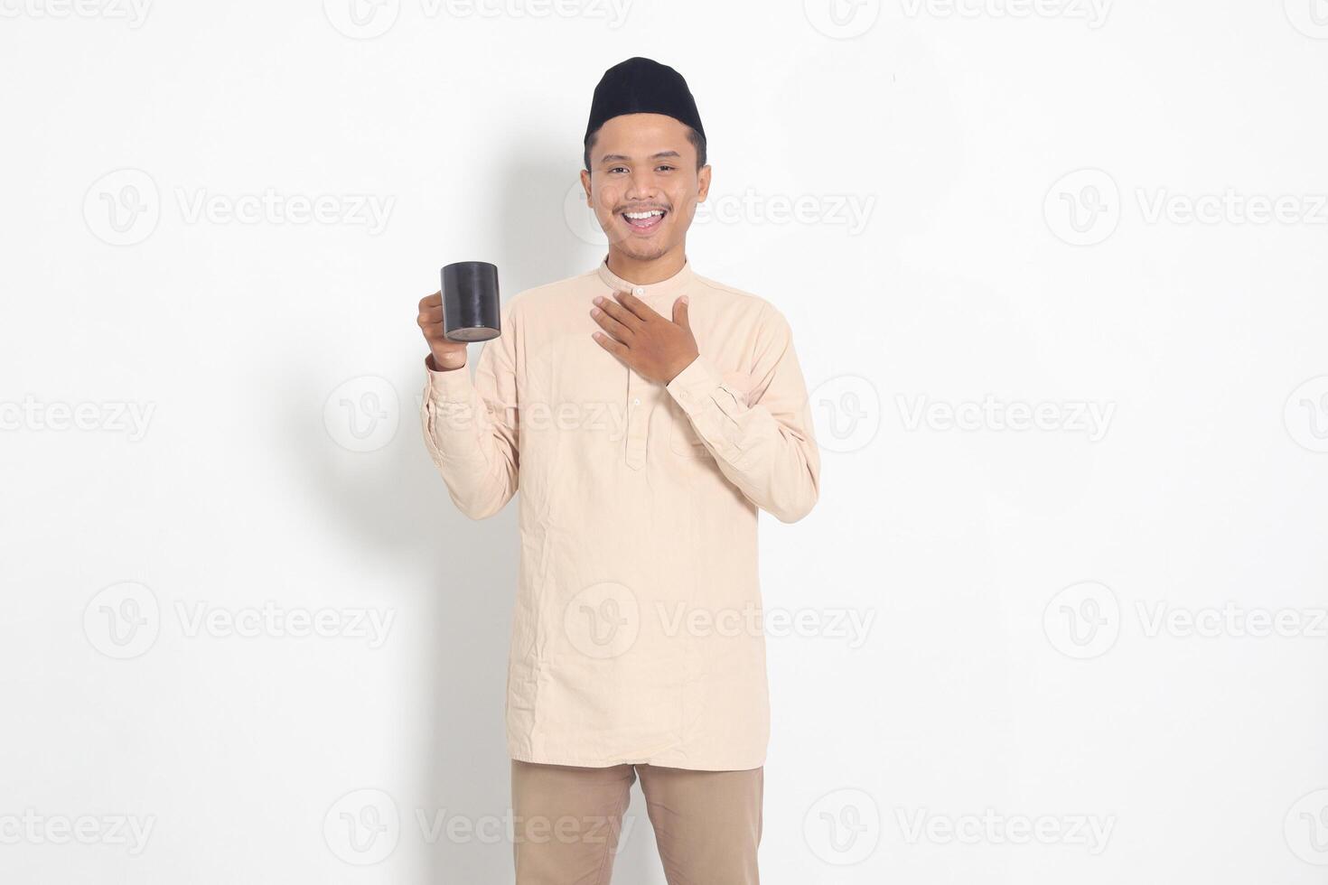 Portrait of attractive Asian muslim man in koko shirt with peci holding a mug, drinking water, quenching thirst after fasting. Isolated image on white background photo
