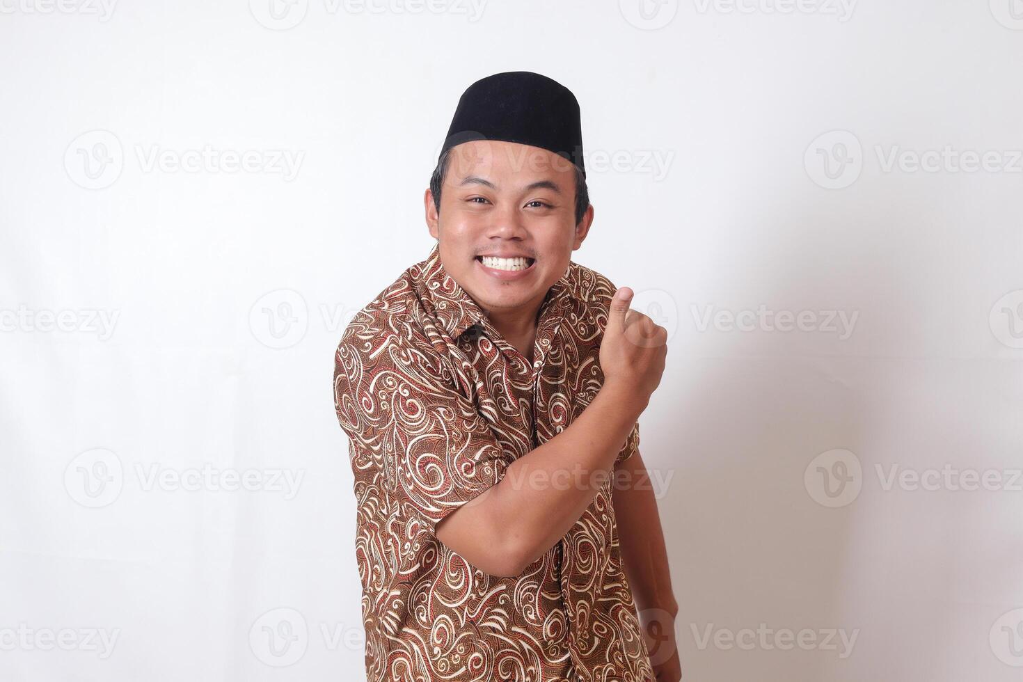 Portrait of excited Asian man wearing batik shirt and songkok smiling and looking at camera, making thumbs up hand gesture. Isolated image on gray background photo
