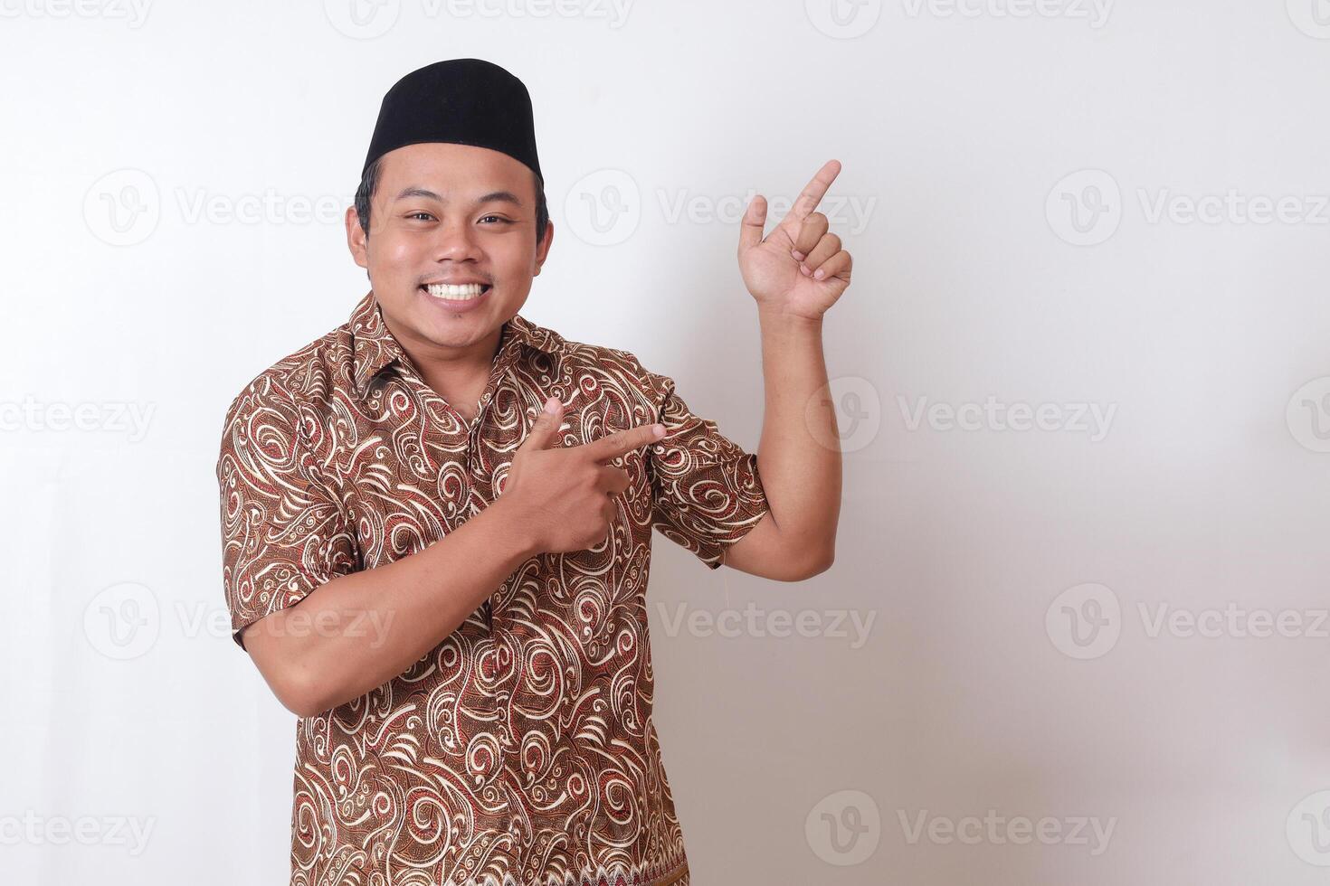 retrato de emocionado asiático hombre vistiendo batik camisa y Songkok sonriente y mirando a el cámara señalando con dos manos y dedos a el lado. aislado imagen en gris antecedentes foto