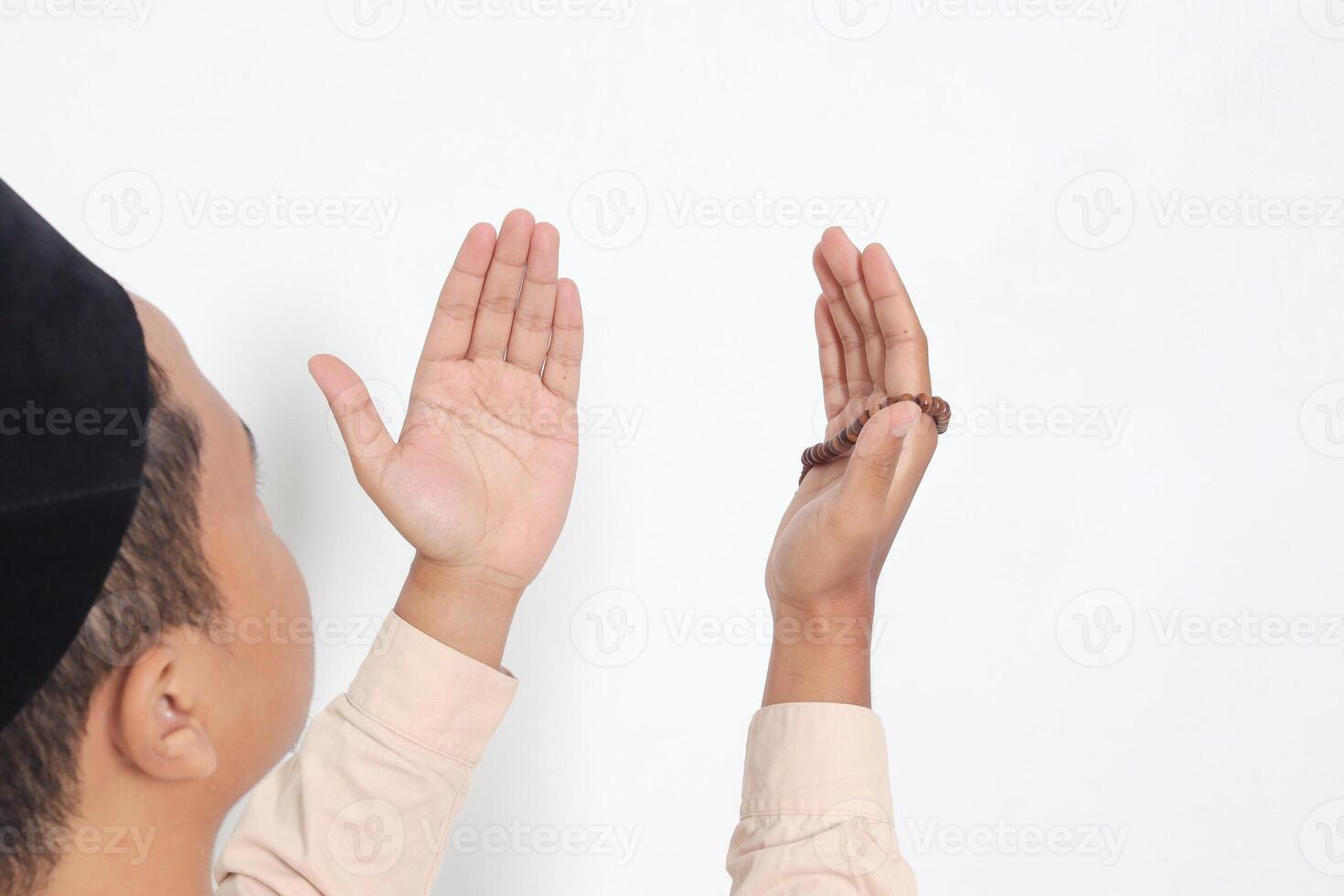 Back view portrait of religious Asian muslim man in koko shirt with skullcap praying earnestly with his hands raised, holding islamic beads. Devout faith concept. Isolated image on white background photo