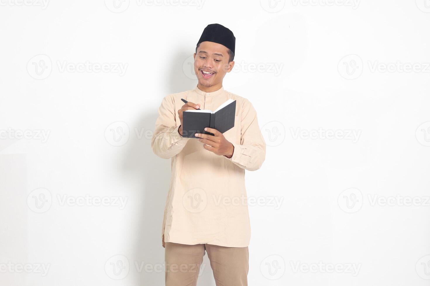 Portrait of excited muslim man in koko shirt with peci writing on his notebook. Happy Asian guy taking notes. Isolated image on white background photo