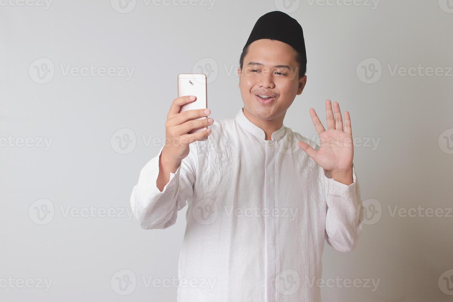 Portrait of Asian muslim man in white koko shirt with skullcap taking picture of himself or selfie, saying hi and waving his hand. Isolated image on gray background photo