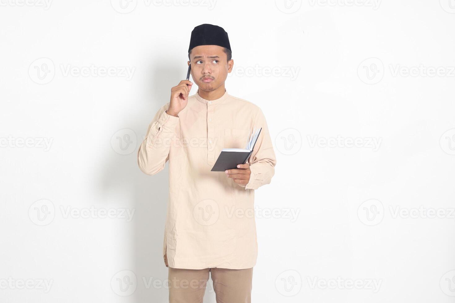 Portrait of confused Asian muslim man in koko shirt with peci reading a book, thinking about an idea with hand holding pen on chin, looking away. Isolated image on white background photo