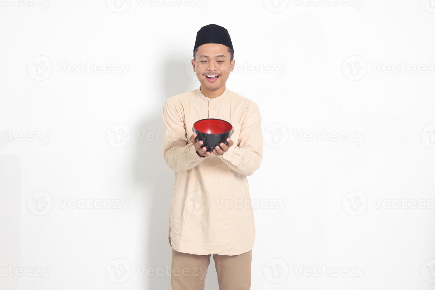Portrait of excited Asian muslim man in koko shirt with skullcap showing and holding an empty plate. Bowl template for food brand. Isolated on white background photo