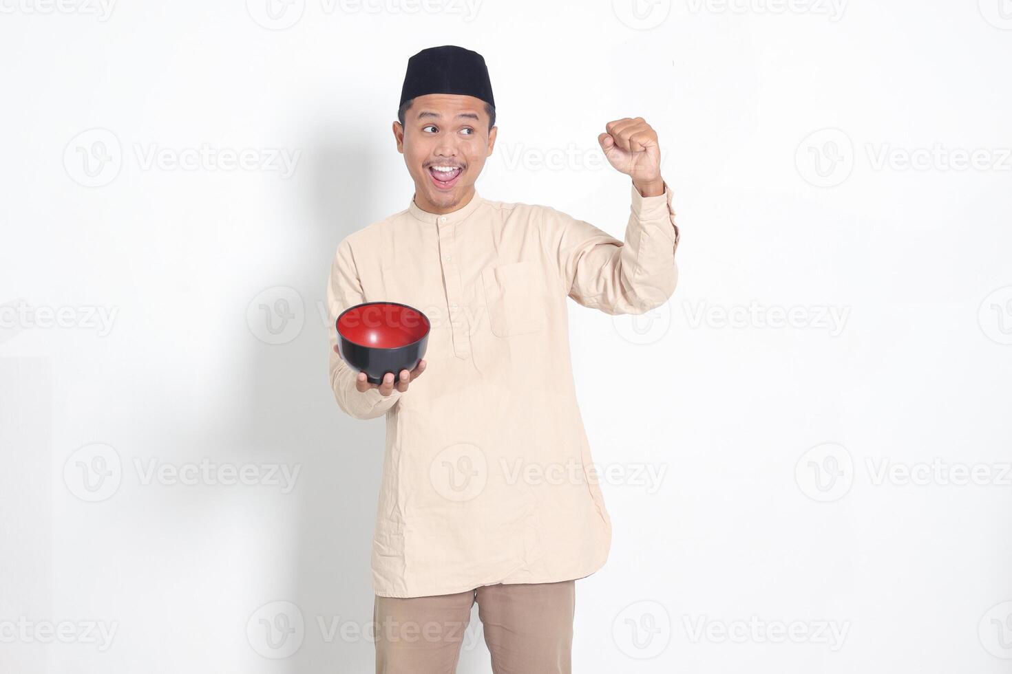 Portrait of excited Asian muslim man in koko shirt with skullcap showing and holding an empty plate. Bowl template for food brand. Isolated on white background photo