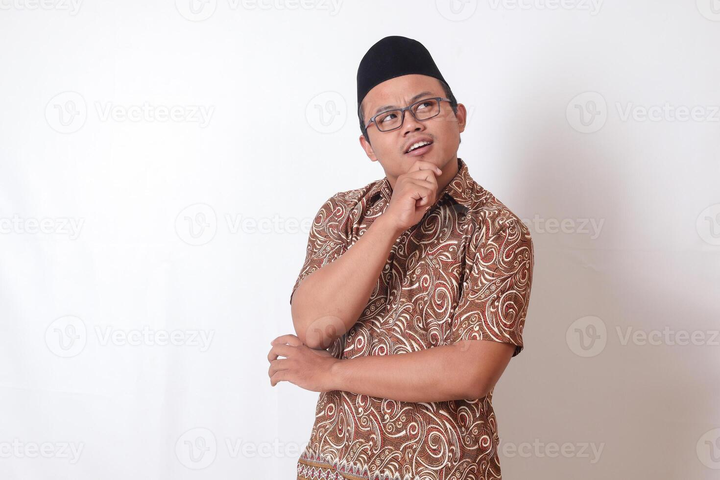 Portrait of confused Asian man wearing batik shirt and songkok standing against gray background, thinking about question with hand on chin photo