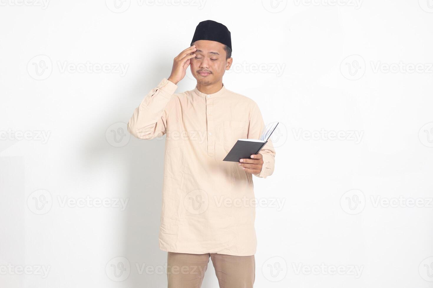 Portrait of confused muslim man in koko shirt with peci reading a notebook. Unhappy Asian guy holding his forehead, having headache. Isolated image on white background photo