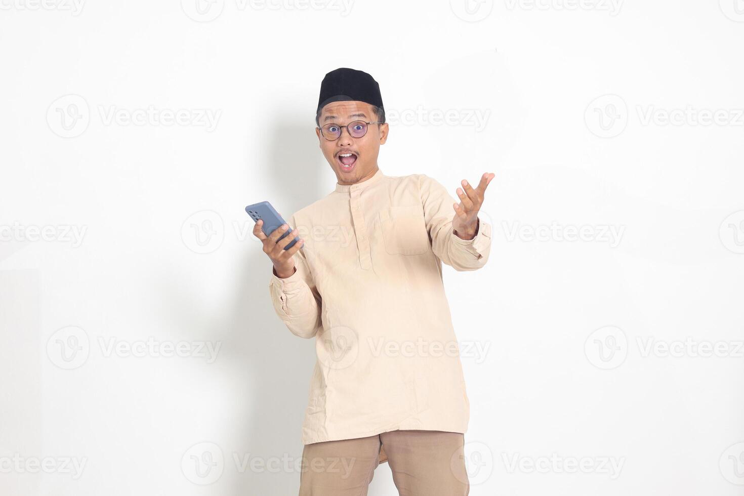 Portrait of surprised Asian muslim man in koko shirt with skullcap holding mobile phone, showing wow shocked face expression. Advertising and social media concept. Isolated image on white background photo