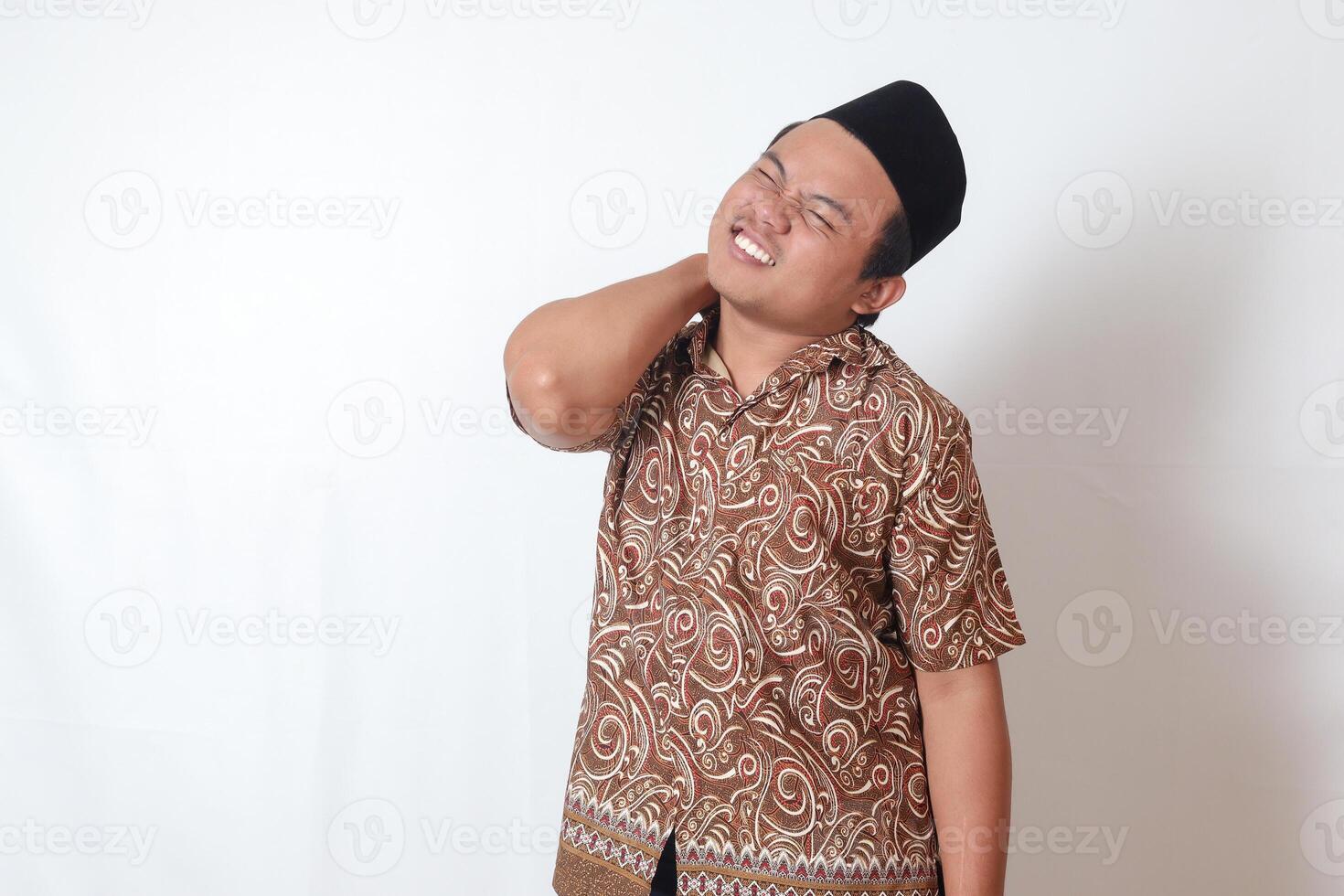Portrait of suffering Asian man wearing batik shirt and songkok having neckache while touching his back. Isolated image on gray background photo