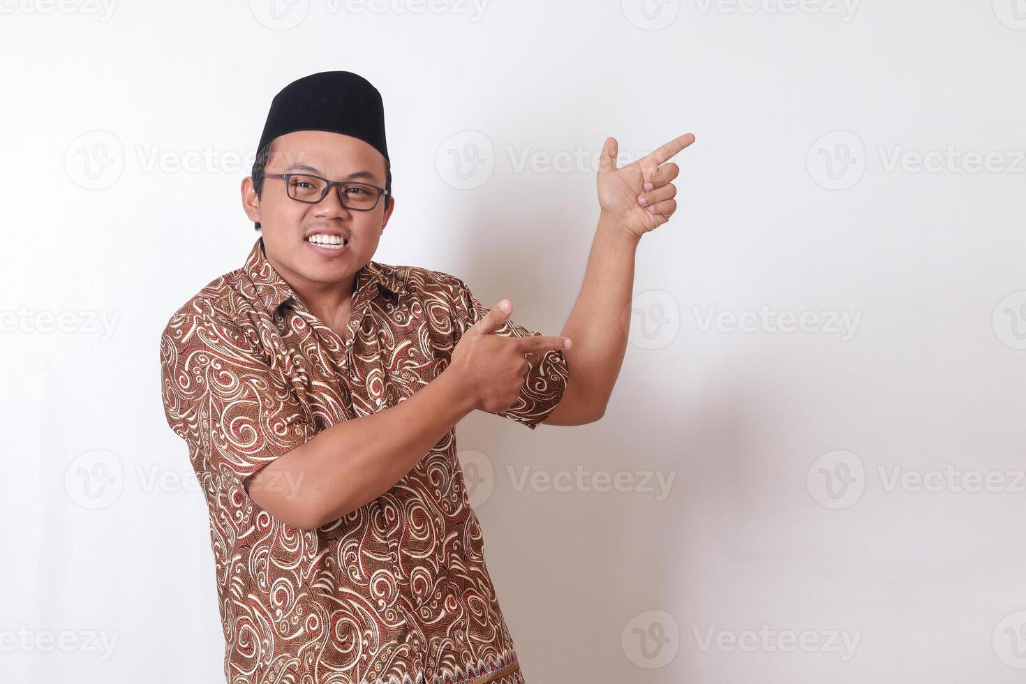 Portrait of excited Asian man wearing batik shirt and songkok smiling and looking at the camera pointing with two hands and fingers to the side. Isolated image on gray background photo