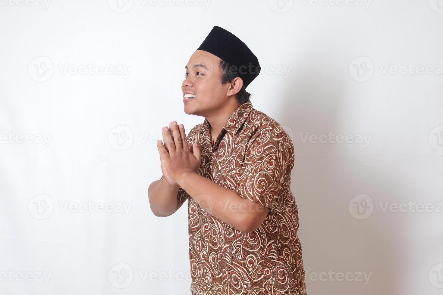 Portrait of smiling Asian man wearing batik shirt and songkok showing apologize and welcome hand gesture. Ramadan advertising concept. Isolated image on gray background photo