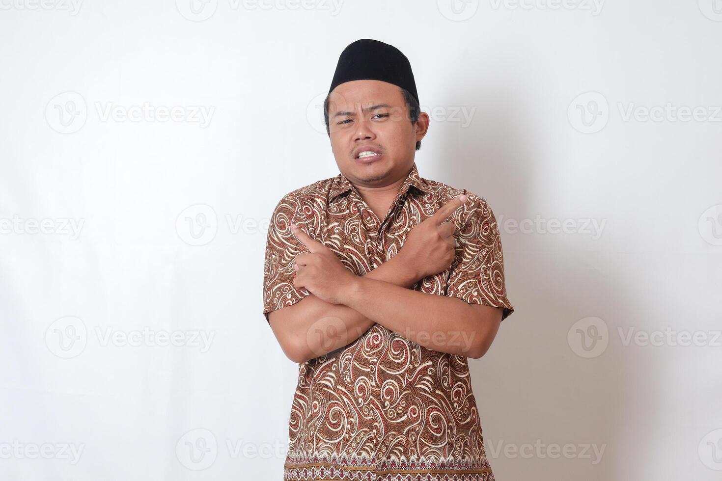 Portrait of confused Asian man wearing batik shirt and songkok with crossed hands, pointing sideways, making choice, choosing between two objects. Isolated image on gray background photo