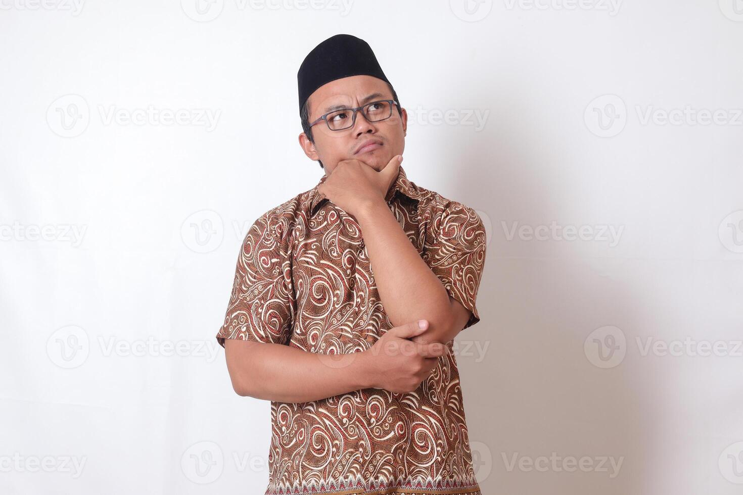 Portrait of confused Asian man wearing batik shirt and songkok standing against gray background, thinking about question with hand on chin photo