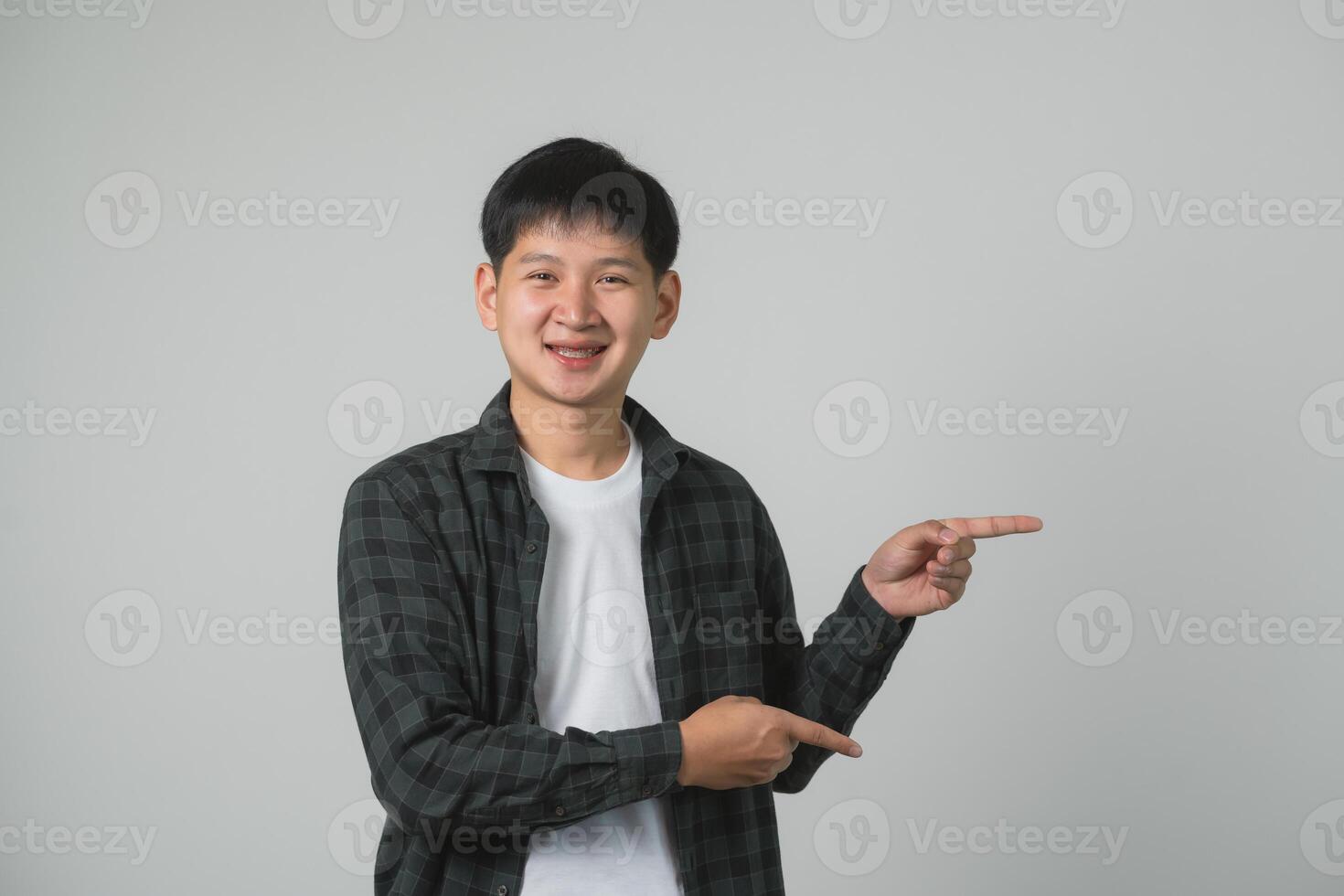 confidente adolescente hombre chico con tirantes señalando hacia arriba. sonriente asiático adolescente en a cuadros camisa señalando hacia arriba en un neutral gris antecedentes. retrato hermoso hombre en estudio concepto. foto