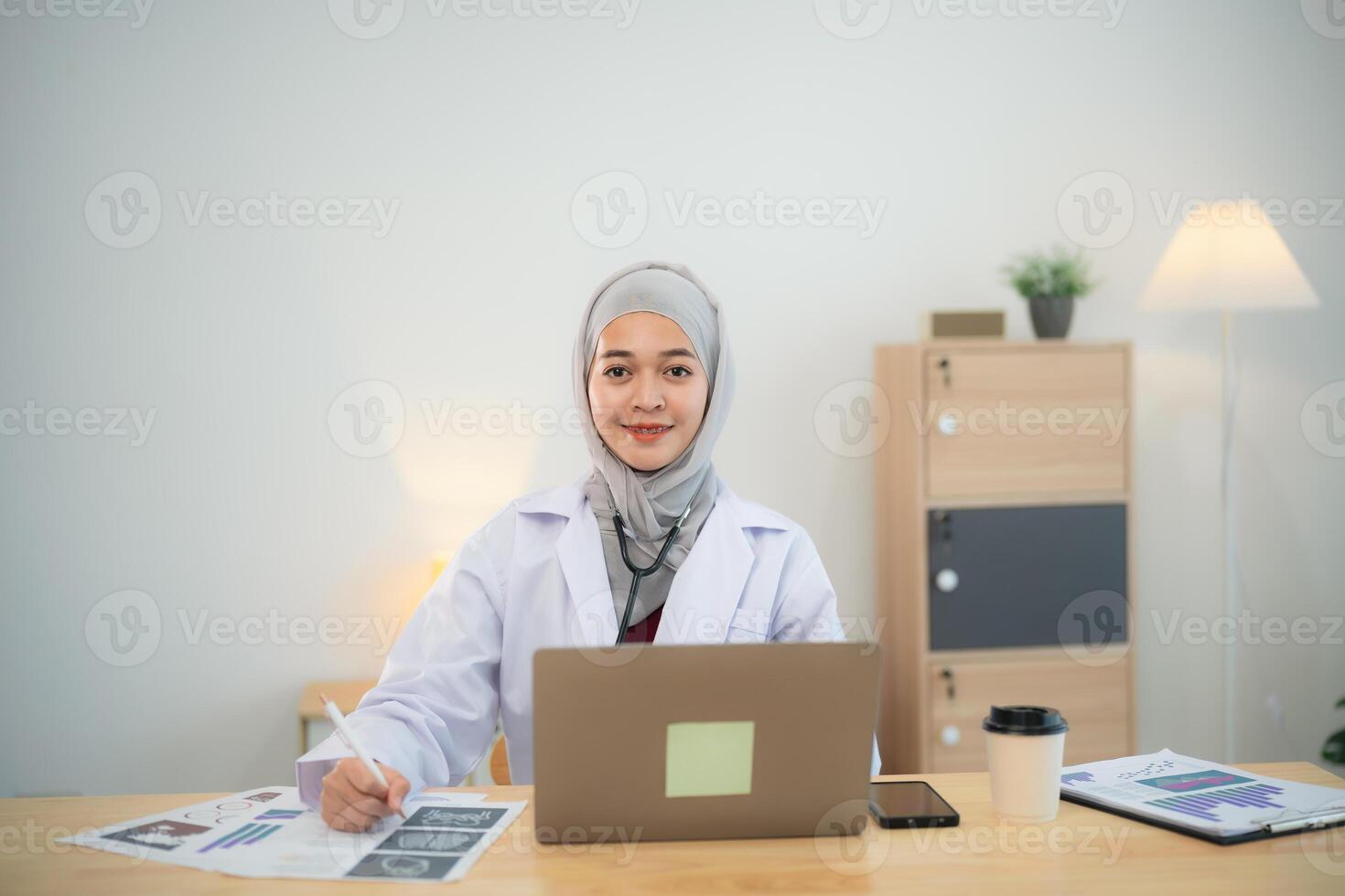 Muslim islam Asian doctor man wearing hijab and stethoscope working with laptop computer and notes on notebook, smartphone. Female doctor work at home office hospital. Health hospital concept. photo