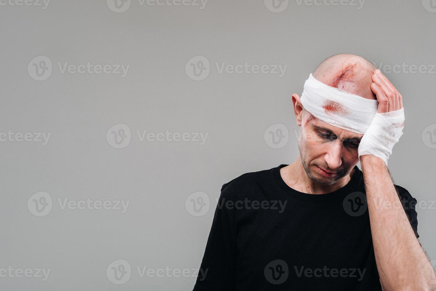 A battered and battered man in a black T-shirt stands against a gray background, holding his aching head with his hands wrapped around it photo