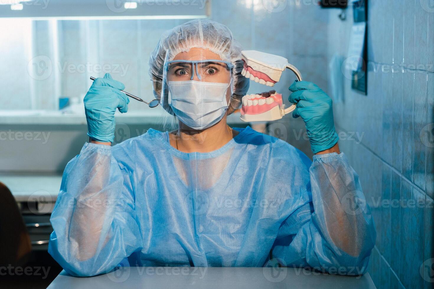 A dental doctor wearing blue gloves and a mask holds a dental model of the upper and lower jaws and a dental mirror photo