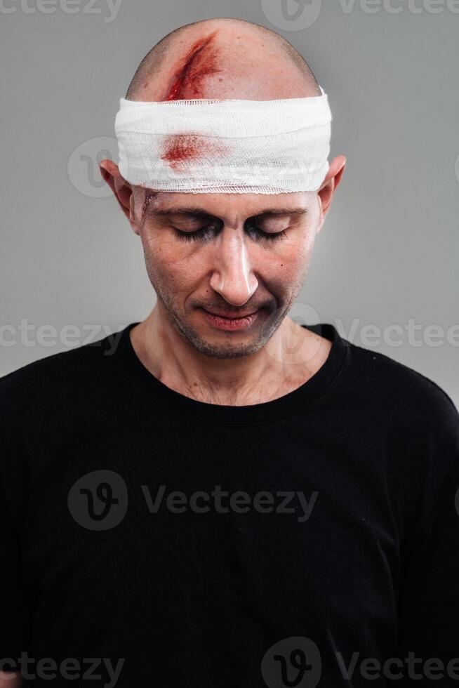 Against a gray background stands a battered and battered man in a black T-shirt with his head wrapped in a bandage photo