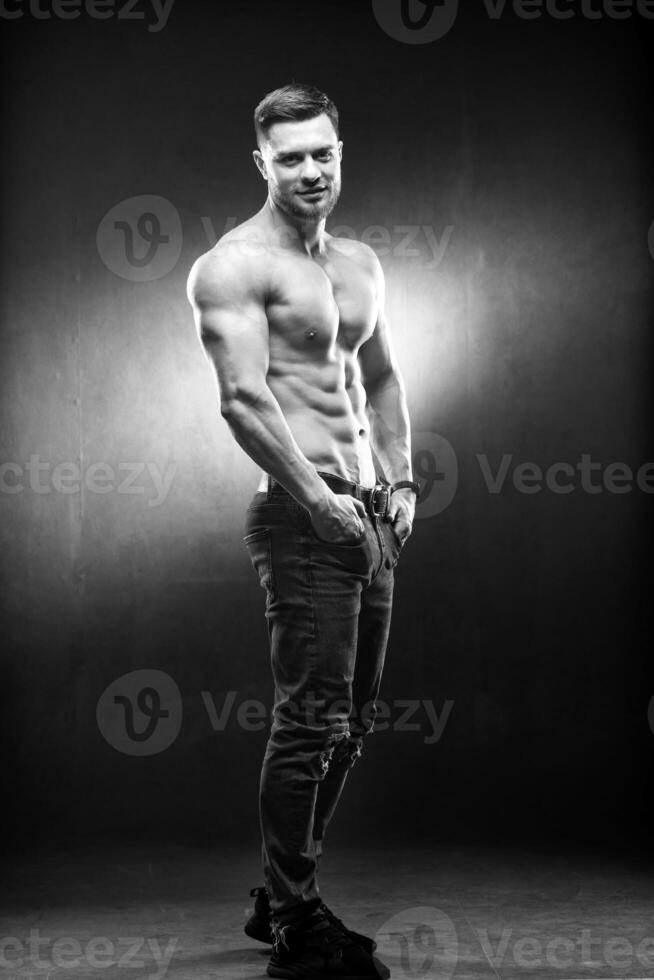 Full size photo of an athletic guy posing with hands in pokets. Studio shot. Bodybuilder. Dark grey background. Closeup.