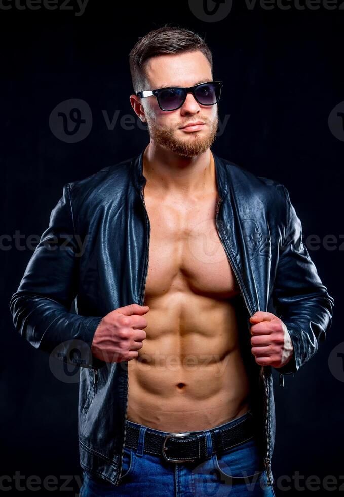 Young handsome macho man in sunglasses with open leather jacket. Man revealing muscular chest and torso. Closeup photo