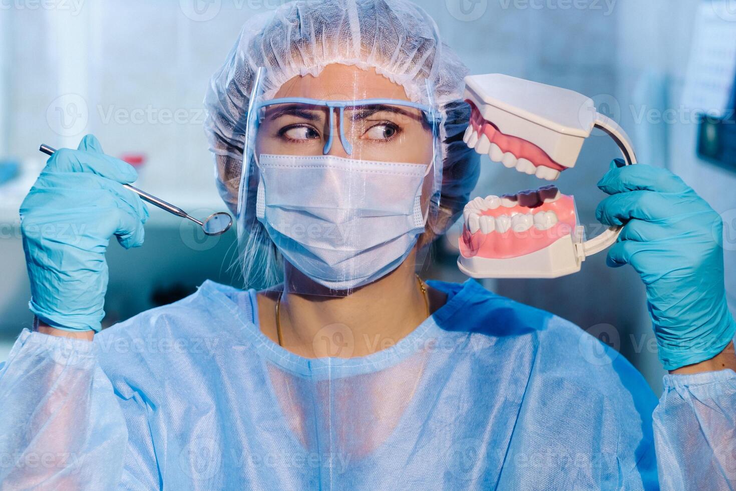 A dental doctor wearing blue gloves and a mask holds a dental model of the upper and lower jaws and a dental mirror photo