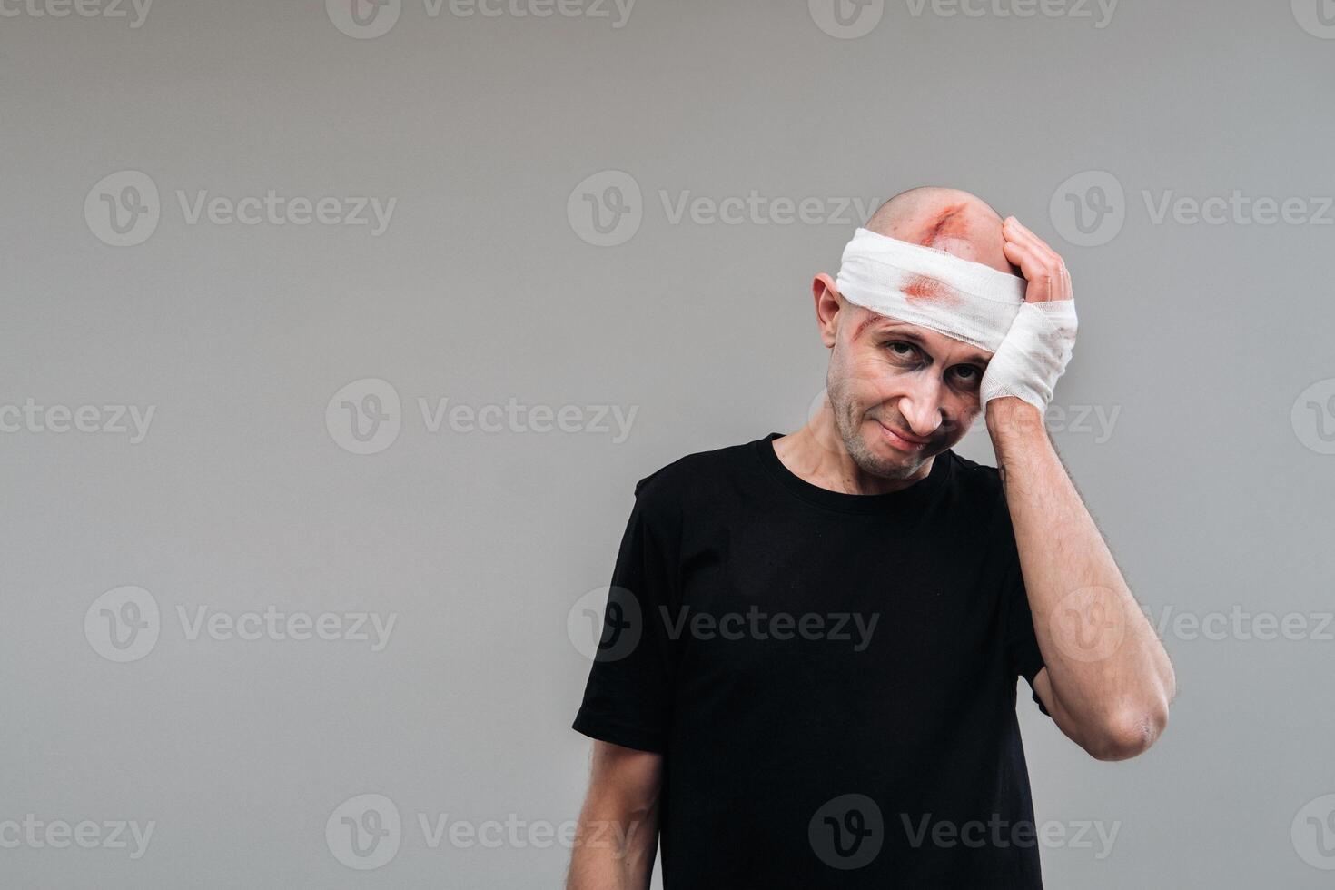 A battered and battered man in a black T-shirt stands against a gray background, holding his aching head with his hands wrapped around it photo