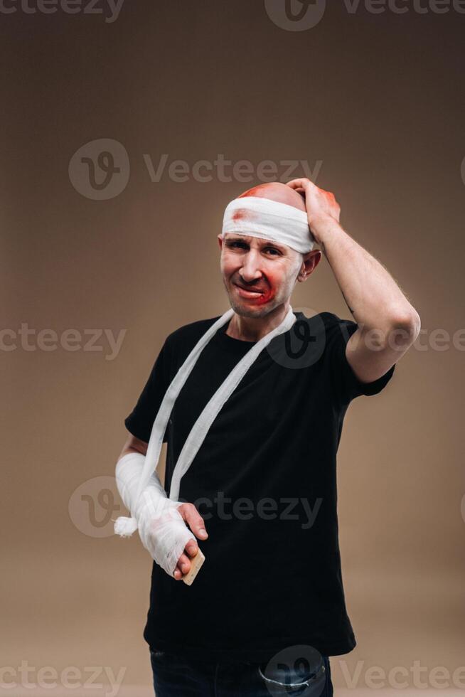 A battered man with a bandaged head and a cast on his arm stands on a gray background photo