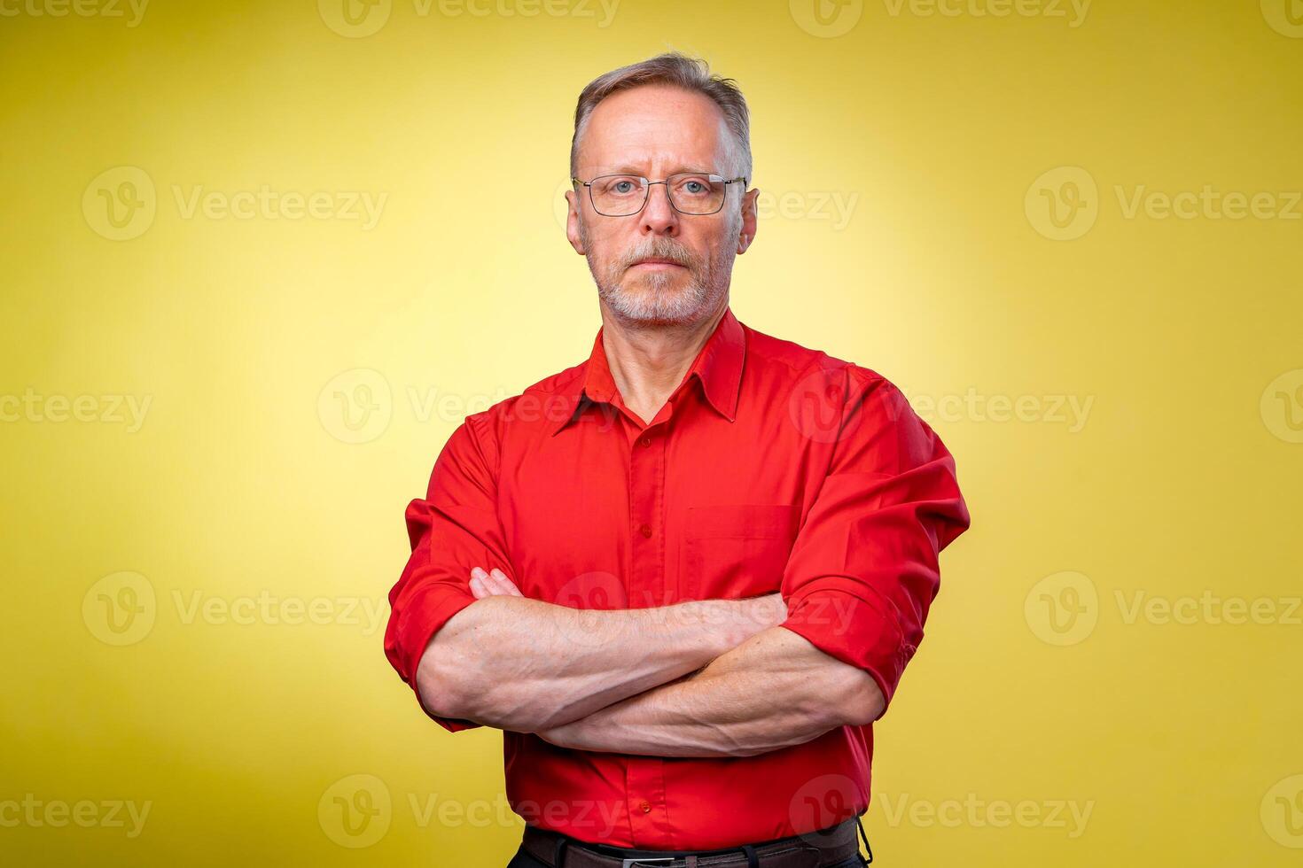 Aged bearded fit man is standing with crossed hands isolated against yellow background. Red shirt. Man in glasses. photo