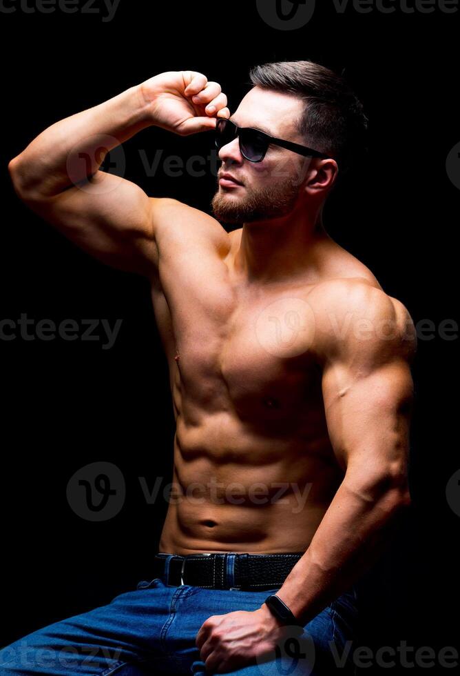 Well build handsome man without a shirt posing in studio. Strong body and cool face. Male wearing glasses, sitting on a chair with hands on legs. Closeup. photo
