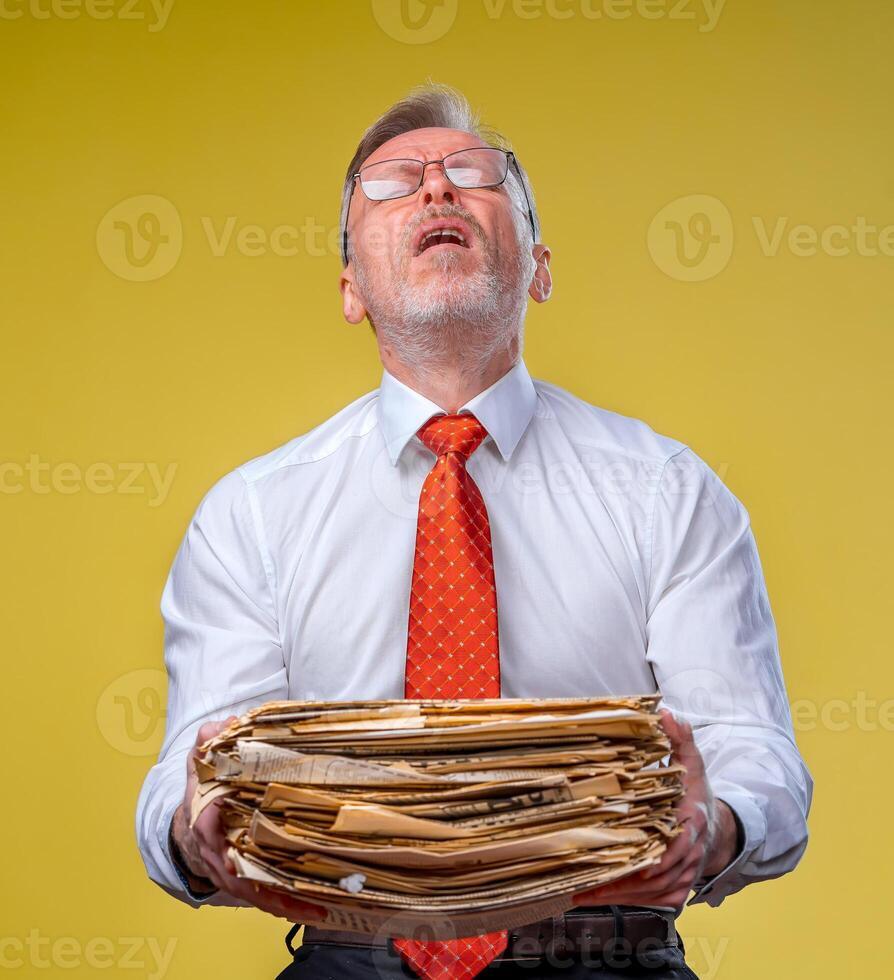 A businessman carrying pile of paperwork. Yellow background. Sad expression. To do a lot photo