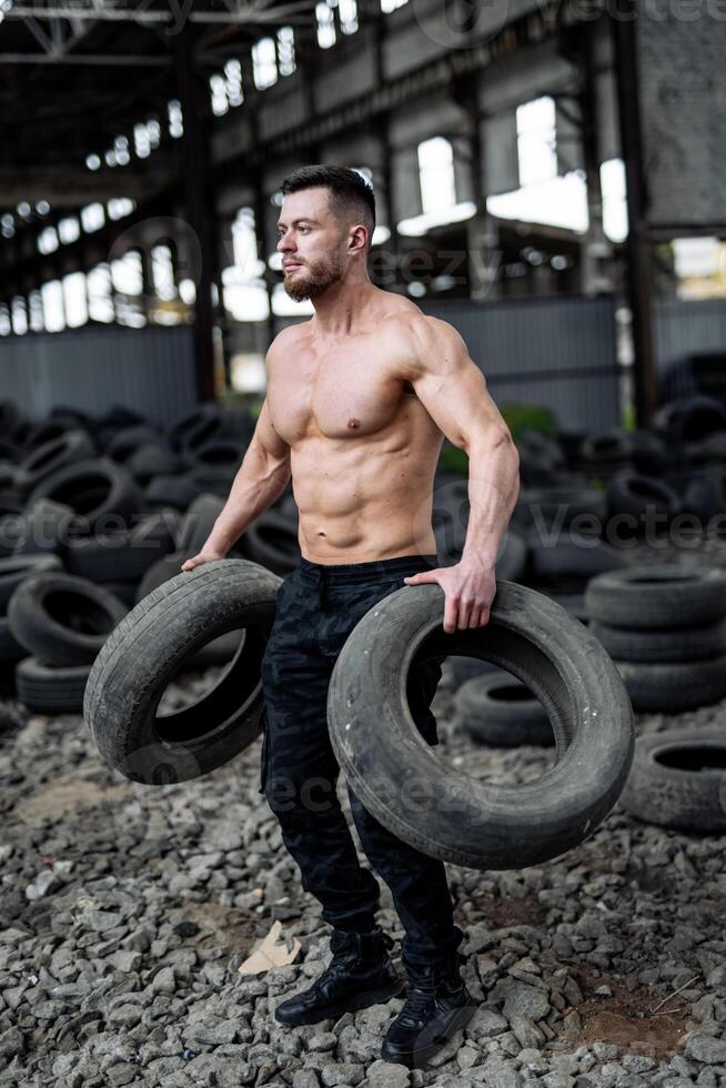 fuerte joven hombre ascensores llantas sin camisa. grande músculos y Perfecto abdominales. muchos llantas en el antecedentes. hombre fuerte tiene rutina de ejercicio foto