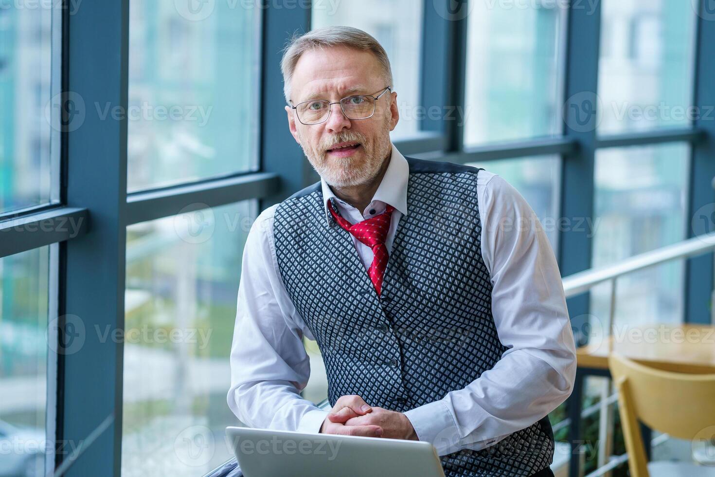 Elderly man is sitting in a light room with big glass windows near computer. Working on a latop in modern restaurant during coffee brake. Looking at camera. photo