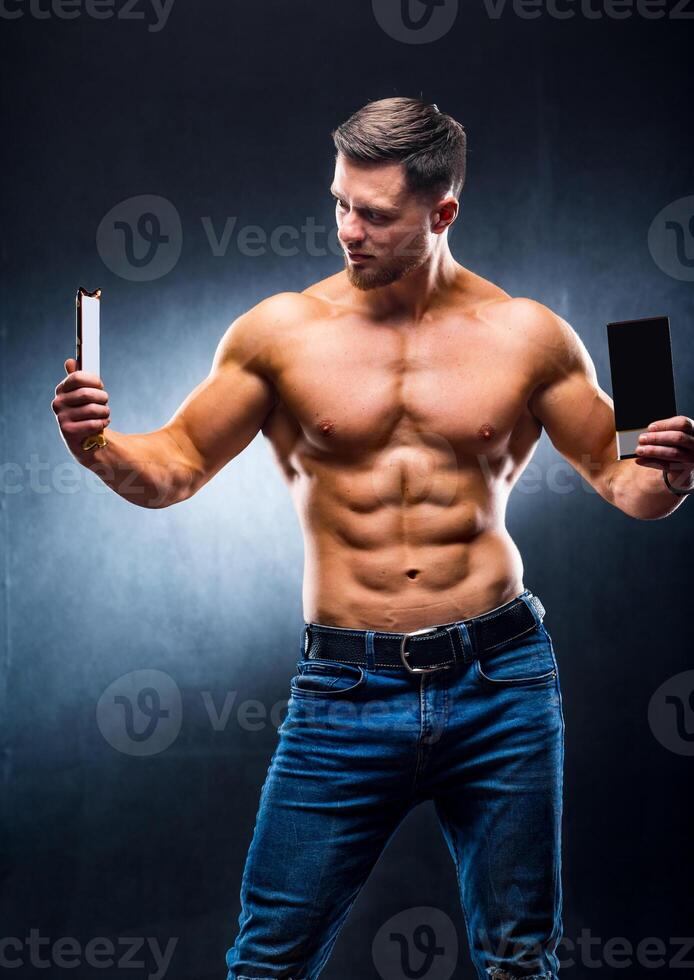 Bodybuilder sportsman holding sport bar and chocolate . Choosing between healthy and harmful food. Naked torso. Grey background. Vertical photo. Closeup photo