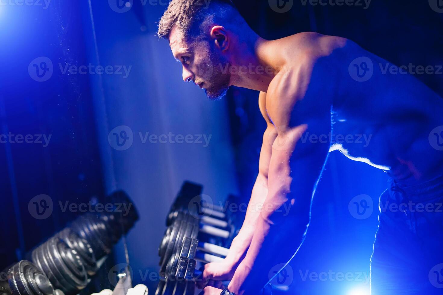 Fit muscular man exercising with dumbbell. Muscular young man lifting weights on dark background. Blue light filter, naked torso. Closeup. photo