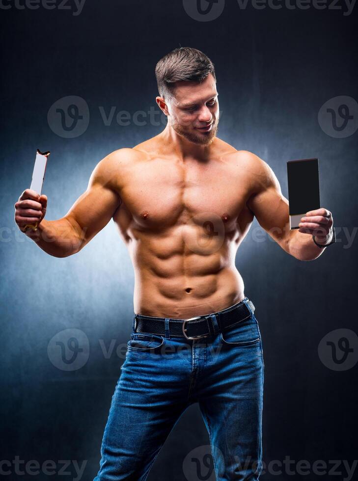 Bodybuilder sportsman holding sport bar and chocolate . Choosing between healthy and harmful food. Naked torso. Grey background. Vertical photo. Closeup photo